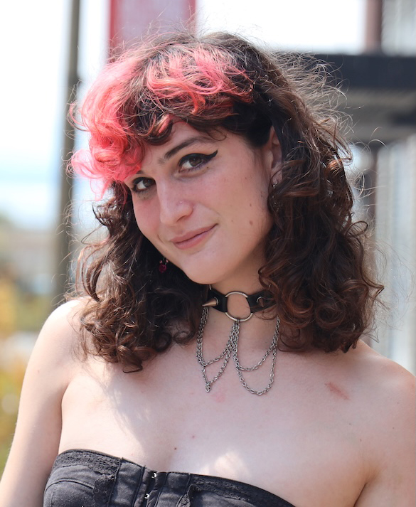 Brunette woman with pink highlights wearing a black dress and a studded choker. She is smiling at you.